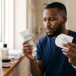African man comparing exfoliating pads and facial scrubs for acne-prone skin during a skincare routine.